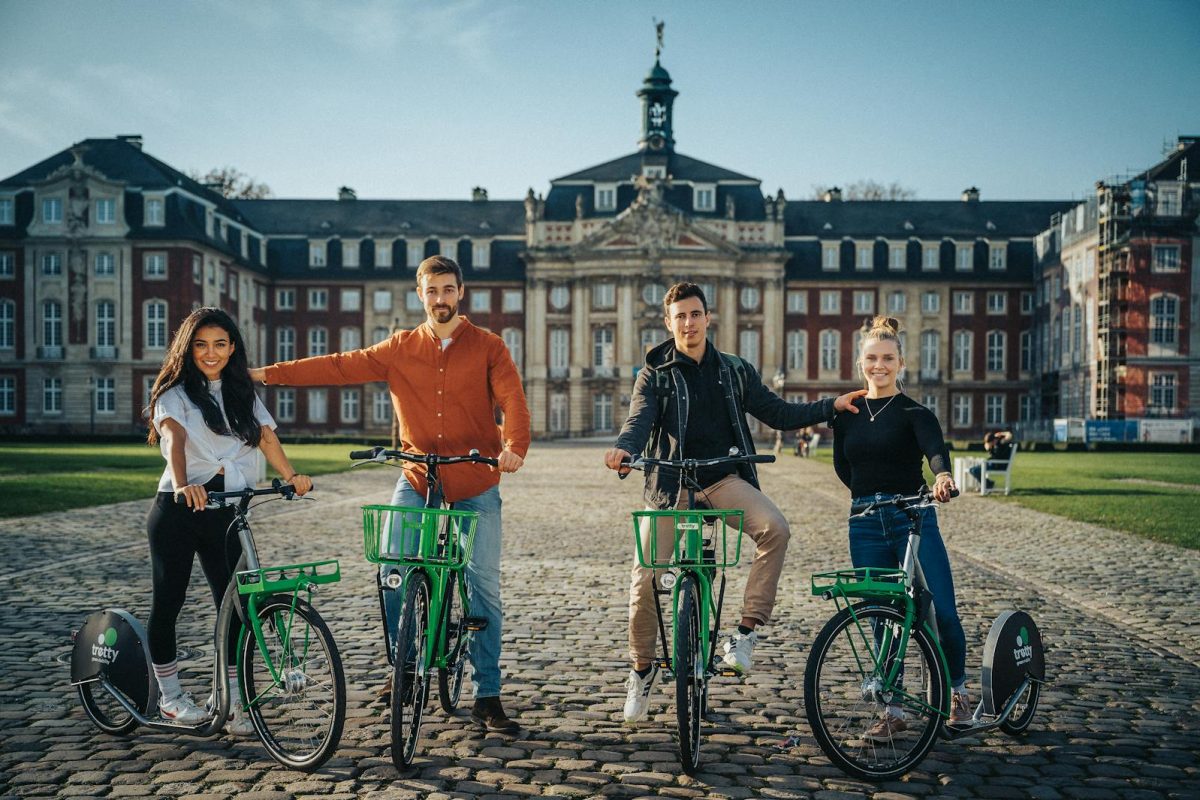 A diverse group of young adults posing with rental bikes in front of a historic building on a sunny day.